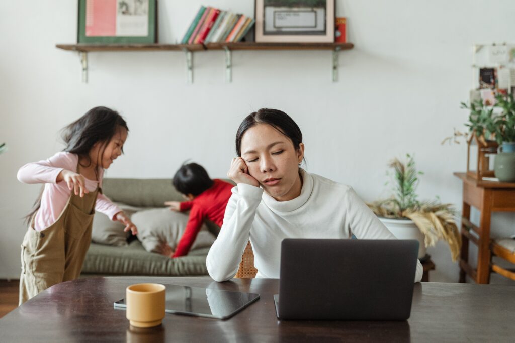 a tired-looking woman works at a desk while two children play in the background